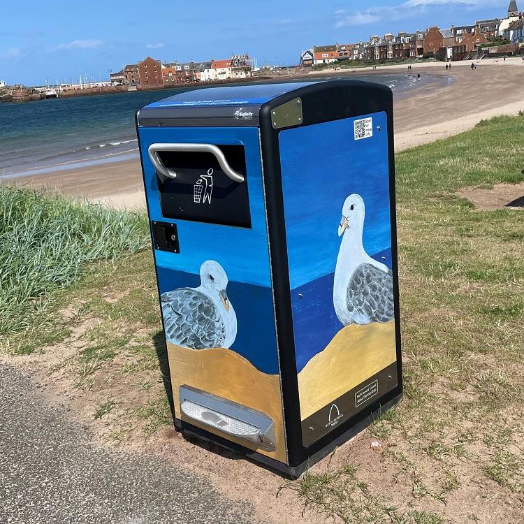 A large bin stands beside a path by the sea, bathed in sunshine, with North Berwick Harbour and West Beach in the background. The bin has a foot peddle at the base for opening the 'trap door' into which waste is discarded. The bin is beautifully and entirely covered in seabird (fulmar) illustrations. A large bin stands beside a path by the sea, bathed in sunshine, with North Berwick Harbour and West Beach in the background. The bin has a foot peddle at the base for opening the 'trap door' into which waste is discarded. The bin is beautifully and entirely covered in seabird (fulmar) illustrations.