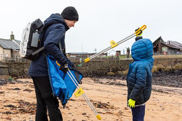 Two people work together to pick up rubbish off the beach, with the Scottish Seaird Centre in the background