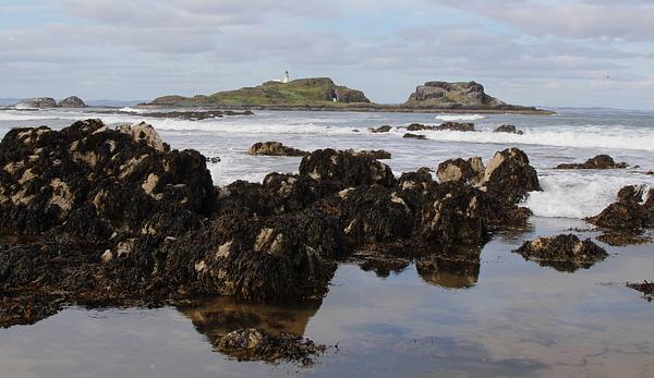 The island of Fidra spans the skyline on an overcast day, with a rocky shore visible in the immediate foreground